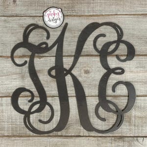 May include: Black metal monogram with the letters "KE" in a decorative script font. The monogram is on a light wood background.
