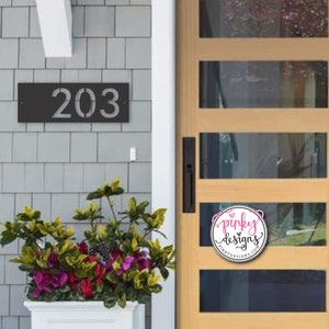 A modern address plaque mounted on a light gray tile of a home. The address sign is painted black and reads 203. To the right of the sign is a modern looking light brown door with windows cut out of it. Below the sign is a planter blooming.