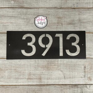 There is a horizontal address sign reading 3913 and is left aligned on the plaque. The metal is painted Oil Rubbed Bronze to showcase one of the finish options. It's laid on a light wood background with a pinky designs logo above.