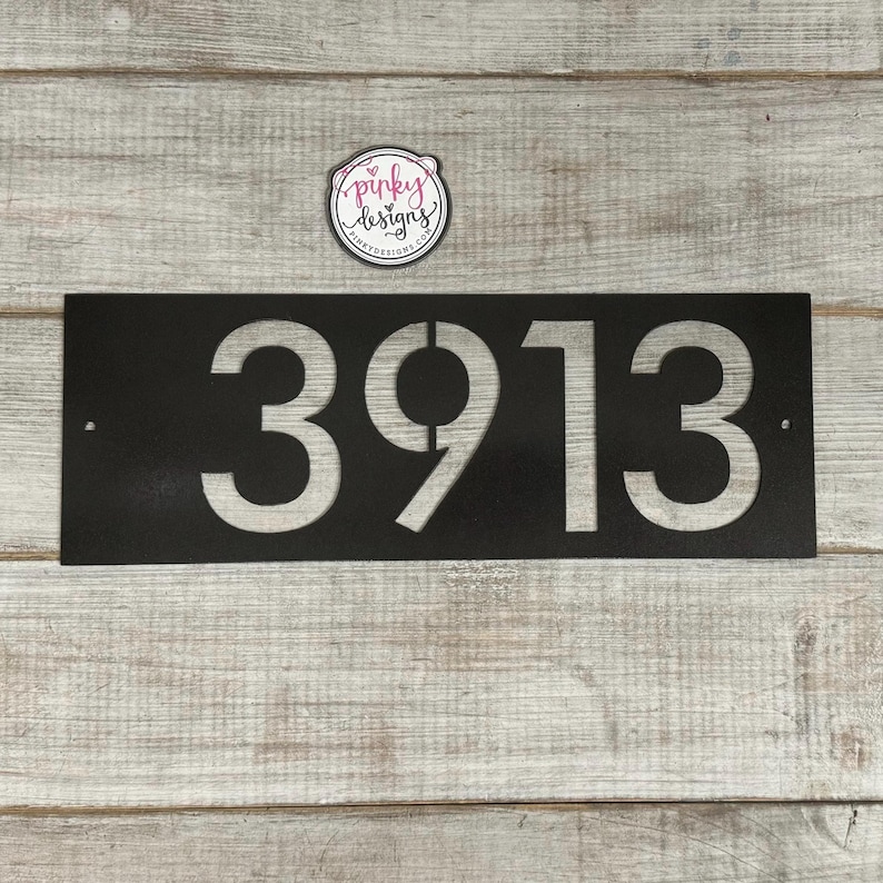There is a horizontal address sign reading 3913 and is left aligned on the plaque. The metal is painted Oil Rubbed Bronze to showcase one of the finish options. It's laid on a light wood background with a pinky designs logo above.
