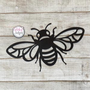 May include: Black metal bee cutout with detailed wings and a striped body. The bee is mounted on a light wood background.