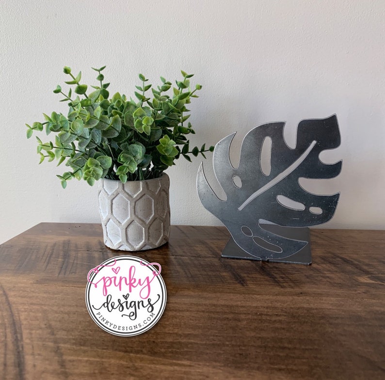 A metal cutout piece thats cut into the shape of a monstera leaf. The sign is bent to be a bookend. There is a plant to the left of the bookends with a Pinky Designs logo below.