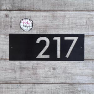 May include: A rectangular black metal house number sign featuring the cut-out digits 217. The sign has two small circular mounting holes on the left and right sides. A circular logo sticker with the text pinky designs is positioned above the sign.