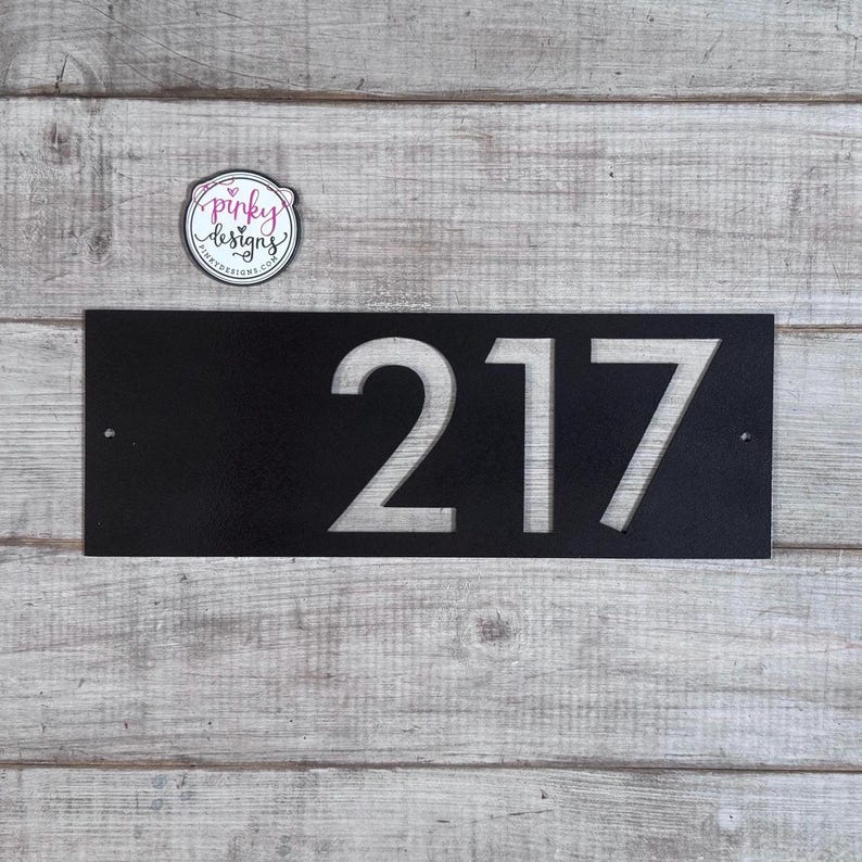 May include: A rectangular black metal house number sign featuring the cut-out digits 217. The sign has two small circular mounting holes on the left and right sides. A circular logo sticker with the text pinky designs is positioned above the sign.