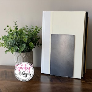 May include: A metal bookend with a gray finish, holding up two books with white and yellow covers. The bookend is on a wooden surface with a small potted plant in the foreground.