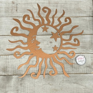 May include: Copper metal wall art featuring a sun with a crescent moon and stars. The sun has a face and curly rays. The piece is on a wood background.