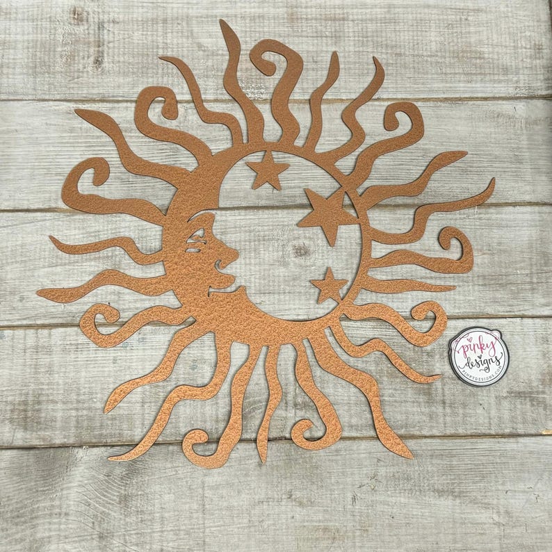 May include: Copper metal wall art featuring a sun with a crescent moon and stars. The sun has a face and curly rays. The piece is on a wood background.