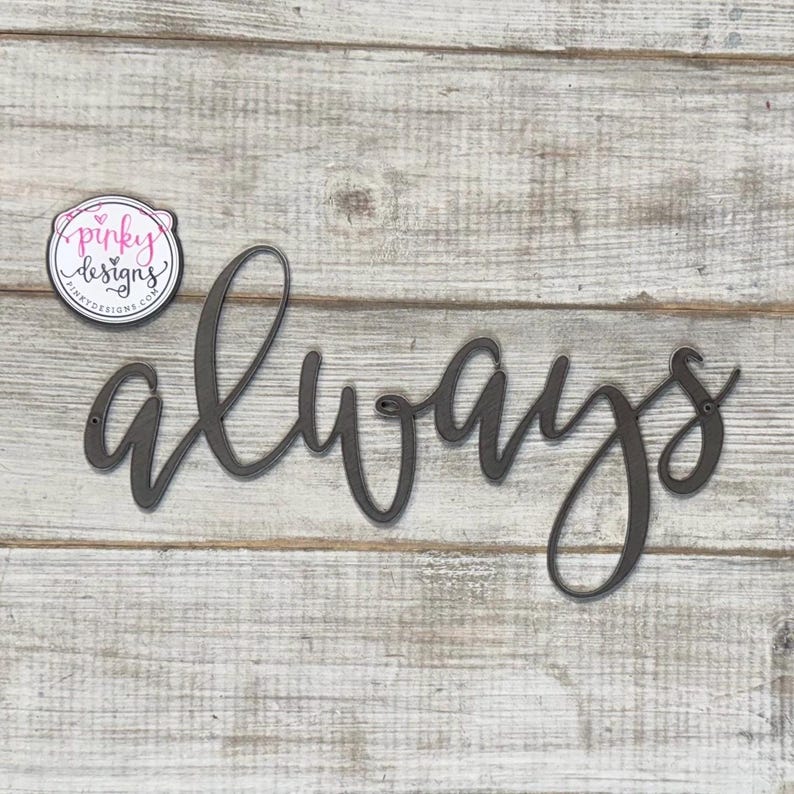 May include: A metal sign with the word "always" written in a cursive font. The sign is painted a dark gray color and is mounted on a white wooden background.