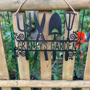 A metal gardening decor piece with 4 gardening tools, there is a line in between them for the wording. The sign reads Grampys Garden and is painted black and mounted on a wooden fence with a rope. Between the holes in the fence you can see a garden.
