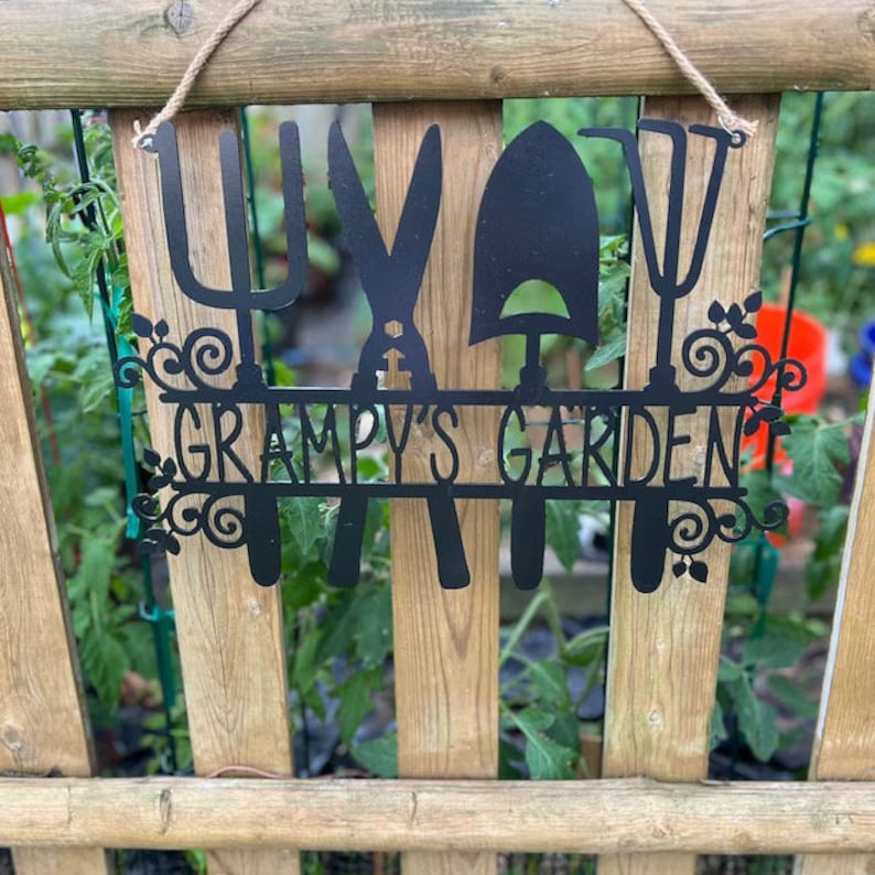 A metal gardening decor piece with 4 gardening tools, there is a line in between them for the wording. The sign reads Grampys Garden and is painted black and mounted on a wooden fence with a rope. Between the holes in the fence you can see a garden.