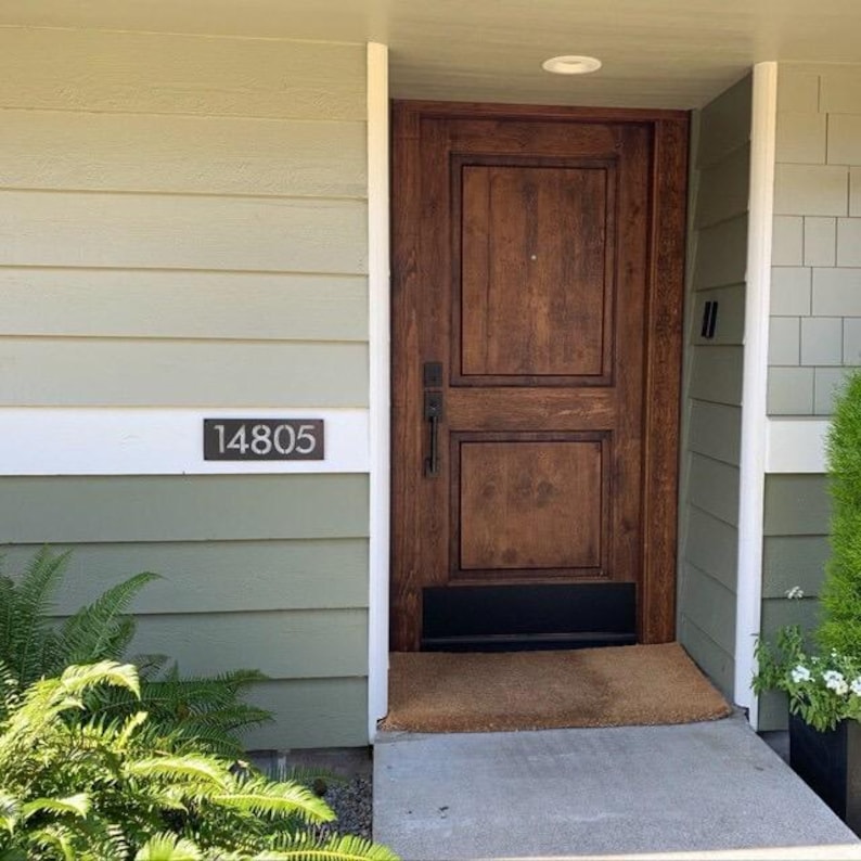 There is a modern rectangular house number sign. The address sign is left raw metal and reads 14805. The sign is mounted on a light green exterior of a home on the only piece that's painted white.