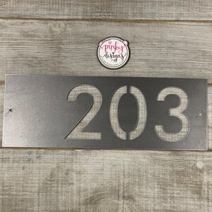 There is a horizontal address sign reading 203 and is left aligned on the plaque. The metal is left Raw Metal really show casing the details of the raw metal patina. It's laid on a light wood background with a pinky designs logo above.