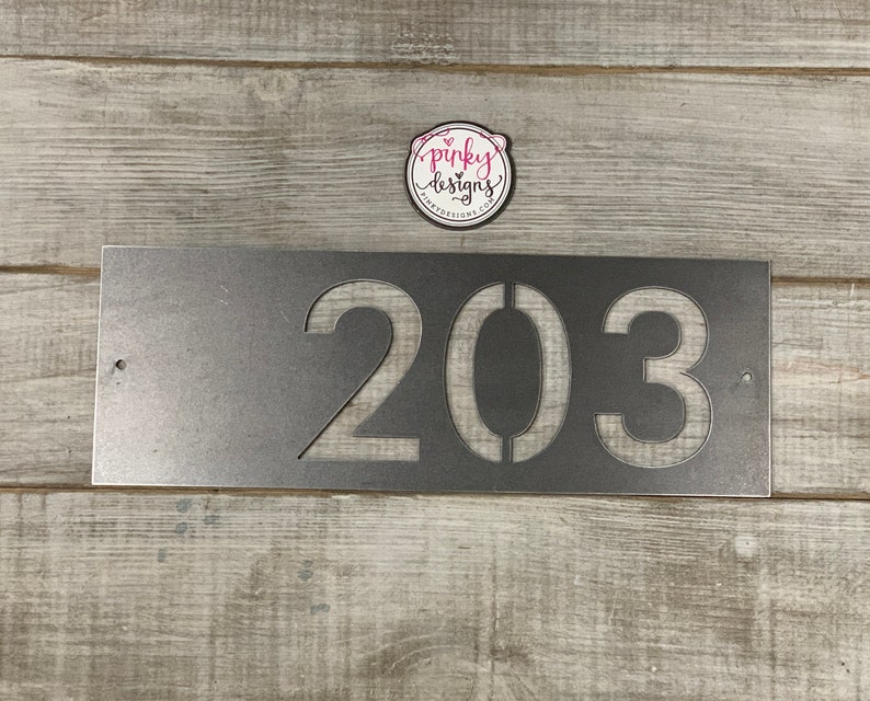 There is a horizontal address sign reading 203 and is left aligned on the plaque. The metal is left Raw Metal really show casing the details of the raw metal patina. It's laid on a light wood background with a pinky designs logo above.