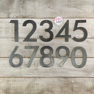 May include: Set of ten metal house numbers, painted in a silver finish. The numbers are arranged in two rows, with the numbers 1 through 5 in the top row and 6 through 0 in the bottom row. The numbers are on a light wood background.
