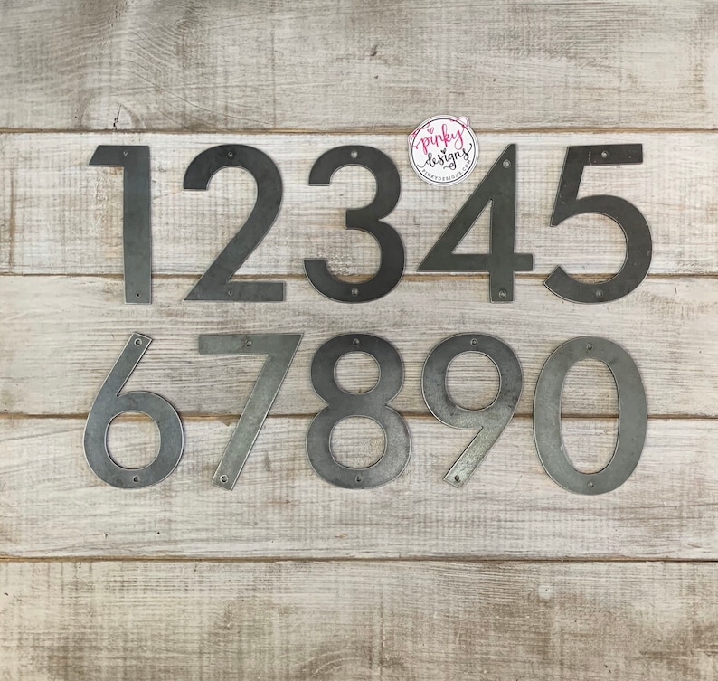May include: Set of ten metal house numbers, painted in a silver finish. The numbers are arranged in two rows, with the numbers 1 through 5 in the top row and 6 through 0 in the bottom row. The numbers are on a light wood background.