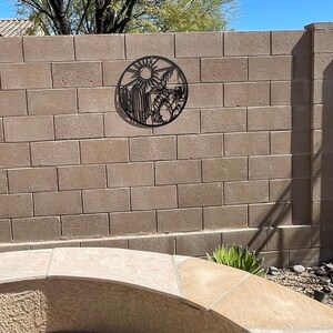 May include: Black metal wall art depicting a desert scene with a sun, cacti, and mountains. The art is mounted on a beige brick wall.