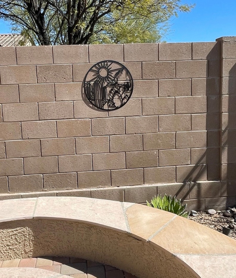 May include: Black metal wall art depicting a desert scene with a sun, cacti, and mountains. The art is mounted on a beige brick wall.