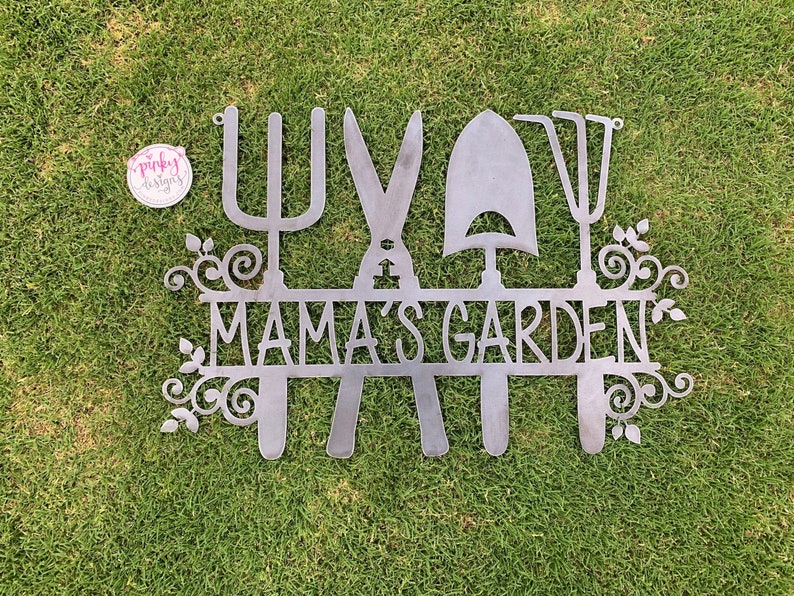 A metal gardening decor piece with 4 gardening tools, there is a line in between them for the wording. The sign reads Mamas Garden and is left raw metal. The sign is laid on green grass to show contrast. There is a pinky designs logo in the upper lef