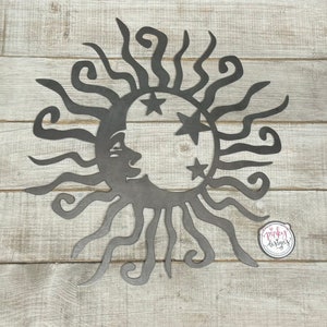 May include: A metal wall decor featuring a sun with a crescent moon and stars. The sun has a face and the moon has a face. The design is in black metal and is on a white wood background.
