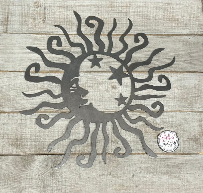 May include: A metal wall decor featuring a sun with a crescent moon and stars. The sun has a face and the moon has a face. The design is in black metal and is on a white wood background.