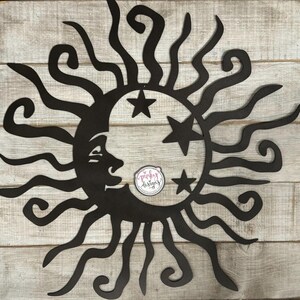May include: Black metal wall art featuring a sun with a crescent moon and stars. The sun has a stylized design with curved rays. The moon has a face and is partially obscured by the sun. The stars are five-pointed.