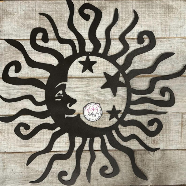 May include: Black metal wall art featuring a sun with a crescent moon and stars. The sun has a stylized design with curved rays. The moon has a face and is partially obscured by the sun. The stars are five-pointed.