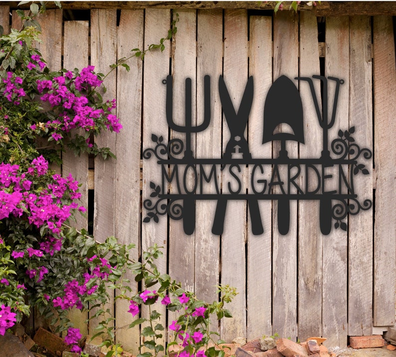 A large metal gardening decor piece with 4 gardening tools, there is a line in between them for the wording. The sign reads Moms Garden and is painted black and mounted on a wooden fence. To the left of the sign is a blooming bush with purple leaves.