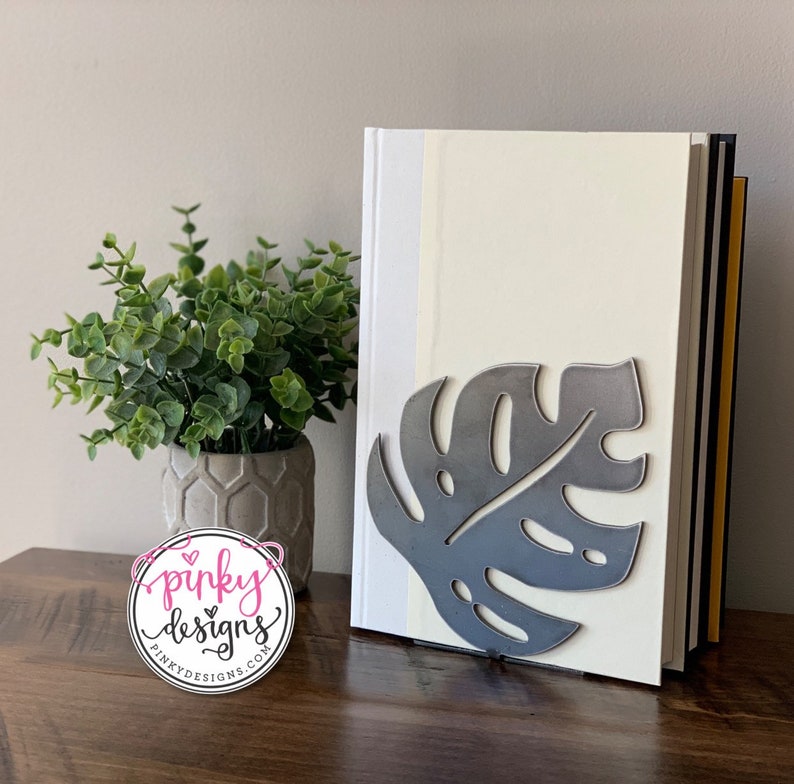 A metal cutout piece thats cut into the shape of a monstera leaf. The sign is bent to be a bookend and is supporting a stack of books. There is a plant to the left of the bookends with a Pinky Designs logo below.