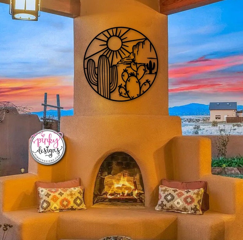 May include: Black metal wall art featuring a desert scene with a sun, mountains, and cacti. The art is hanging on a tan wall above a fireplace.