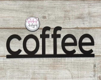 May include: Black metal sign with the word "coffee" in large, stylized letters. The sign is mounted on a light wood background.