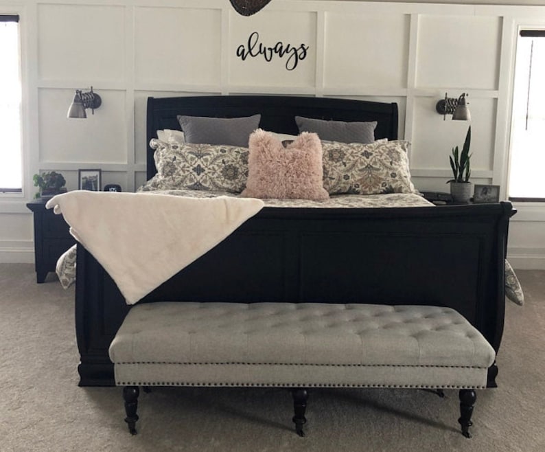 A large metal sign cut out to read the word always. The sign is painted black and mounted above a modern looking headboard. The bed is made with decorative pillows and below is a gray ottoman. The room is very sleek and clean looking.