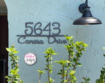 Puede incluir: Un letrero de metal oscuro con el número de casa 5643 y el texto Canosa Drive en letra cursiva. El letrero está montado en una pared azul claro texturizada junto a una lámpara de pared exterior negra y un logotipo circular de Pinky Designs.
