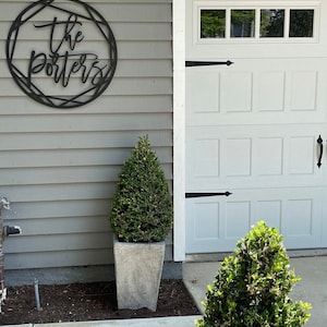 Wall art hung on the exterior of the home on light gray slats. The sign is painted black and reads The Porters. The sign is hung outside next to a white garage below is a concrete planter with a small plant inside it. The home has a modern look.