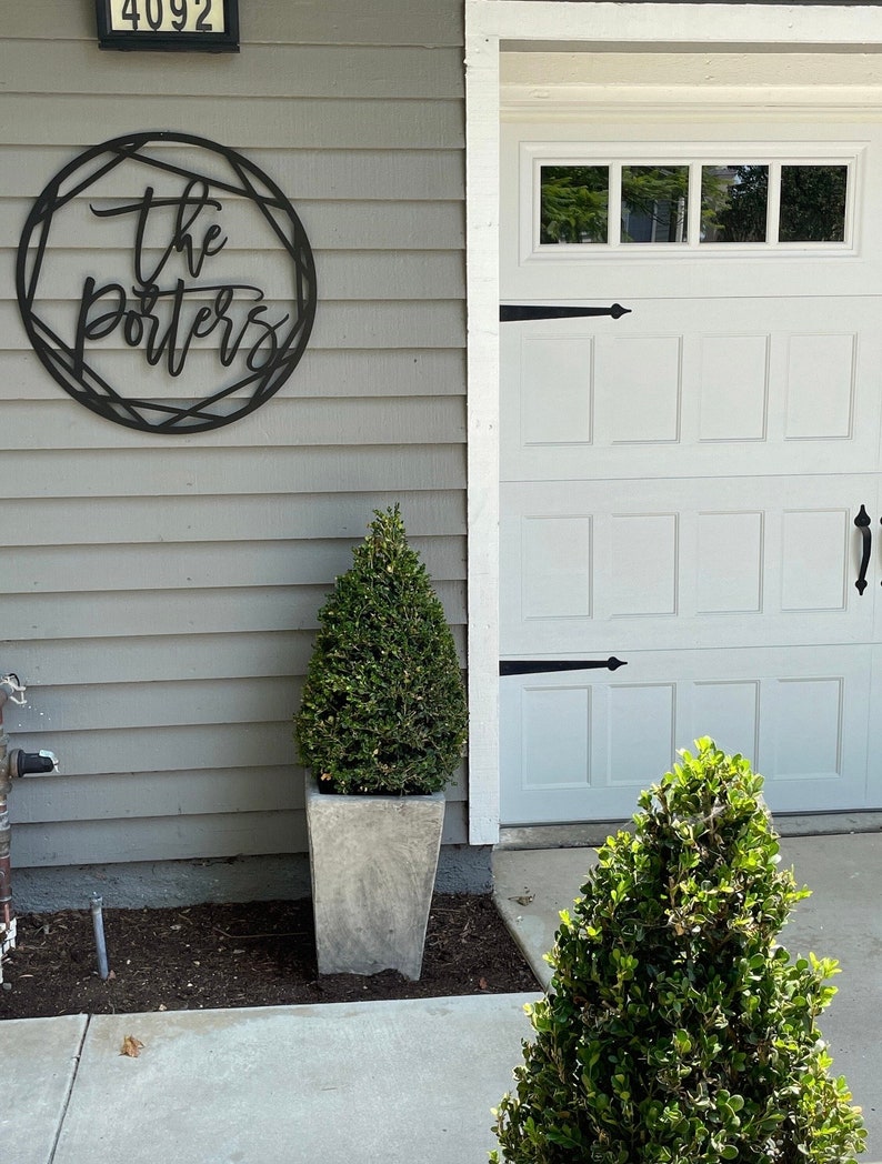 Wall art hung on the exterior of the home on light gray slats. The sign is painted black and reads The Porters. The sign is hung outside next to a white garage below is a concrete planter with a small plant inside it. The home has a modern look.