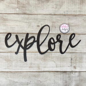 May include: Black metal wall decor with the word "explore" written in a cursive font. The decor is on a white wood background.
