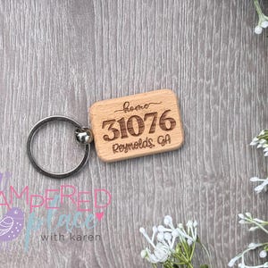 May include: A rectangular light brown wooden keychain with a silver metal ring. The wood is engraved with the word home, the number 31076, and the text Reynolds, GA. The keychain has rounded corners and a smooth, natural wood grain finish.