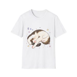 May include: A white short-sleeved t-shirt featuring a graphic illustration of a brown and white cat curled up and sleeping. The cat is surrounded by small colorful stars and blue Zzz text, indicating sleep, centered on the front of the shirt.