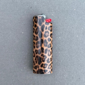 May include: A rectangular pocket lighter featuring a brown and black leopard print pattern across its entire surface. The top section includes a metallic ignition wheel and a small, bright red plastic button for operating the flame mechanism.