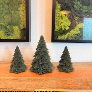 May include: Three conical evergreen tree figurines with a textured, needle-like surface in a deep forest green color. The trees are arranged in a row, with the tallest tree positioned in the center, flanked by two smaller trees of equal height.