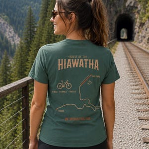 Op de afbeelding: Een donkergroen T-shirt met korte mouwen en een afbeelding op de achterkant met de tekst Route of the Hiawatha, 15 miles, 10 tunnels, 7 trestles en One Unforgettable Ride, inclusief een fietsicoon en een kaart van de route.