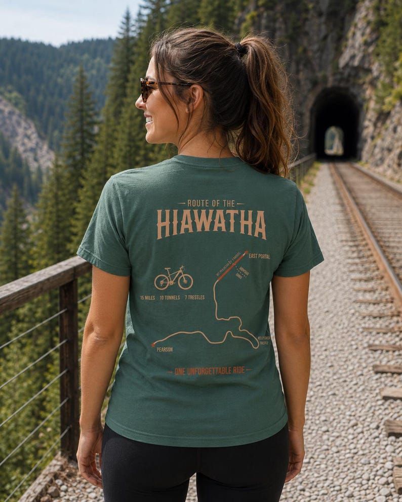 Op de afbeelding: Een donkergroen T-shirt met korte mouwen en een afbeelding op de achterkant met de tekst Route of the Hiawatha, 15 miles, 10 tunnels, 7 trestles en One Unforgettable Ride, inclusief een fietsicoon en een kaart van de route.