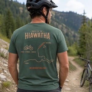 Op de afbeelding: Een bosgroen t-shirt met korte mouwen en een afbeelding op de achterkant met de tekst Route of the Hiawatha en One Unforgettable Ride. Het ontwerp bevat een fietsicoon, een kaart van het pad en labels voor East Portal en Pearson.