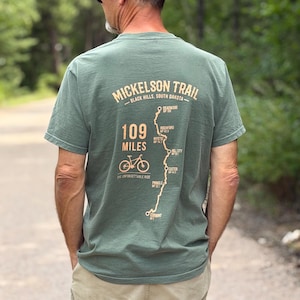 May include: A sage green short-sleeve t-shirt featuring a graphic on the back that reads Mickelson Trail Black Hills South Dakota, 109 Miles, One Unforgettable Ride, with a bicycle icon and a map showing trail locations like Deadwood.