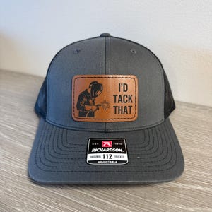 May include: A charcoal grey trucker hat with a black mesh back and a brown leather patch on the front. The patch features a graphic of a welder and the text I'D TACK THAT. A black and white sticker on the brim reads RICHARDSON 112 TRUCKER.