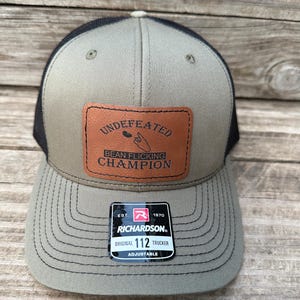 May include: An olive green and black mesh trucker hat featuring a rectangular brown leather patch on the front. The patch displays the text UNDEFEATED BEAN FLICKING CHAMPION with a graphic of a finger flicking a bean. A black sticker is on the brim.