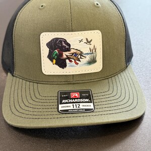 May include: Olive green and black trucker hat with a patch featuring a black Labrador retriever holding a duck. The patch is rectangular with a white background. The hat has the text "RICHARDSON" and "ORIGINAL 112 TRUCKER".
