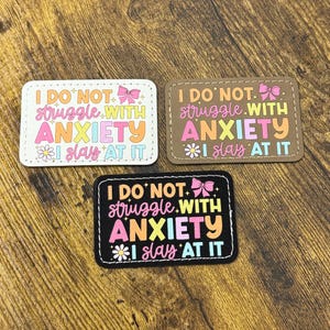 May include: Three rectangular patches in white, brown, and black, each featuring the text I DO NOT struggle WITH ANXIETY I slay AT IT in colorful, stylized lettering. The designs include small pink bows, white daisies, and tiny star accents.