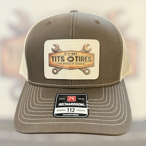 May include: A brown and tan trucker hat with a white patch featuring the text "IF IT HAS TITS OR TIRES I CAN MAKE IT SQUEAL" and two wrenches. The hat has a curved brim and an adjustable strap.