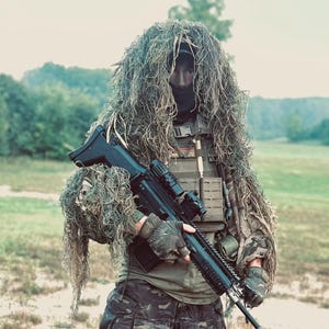 May include: A ghillie suit with long, textured green and brown fibres covering the head and shoulders. The outfit includes a tan tactical vest with a small American flag patch, dark camouflage cargo trousers, and fingerless gloves holding a black rifle.