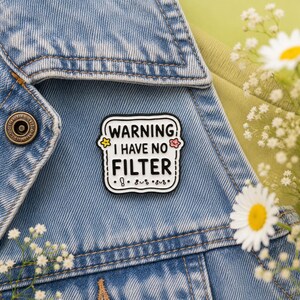 Warning I Have No Filter hard enamel pin displayed on a light blue denim jacket against a soft sage green background with daisy flowers. Funny teacher pin badge, coworker gift, office humour accessory, jacket pin, backpack badge and unique enamel gift by Pocketing Positivity.